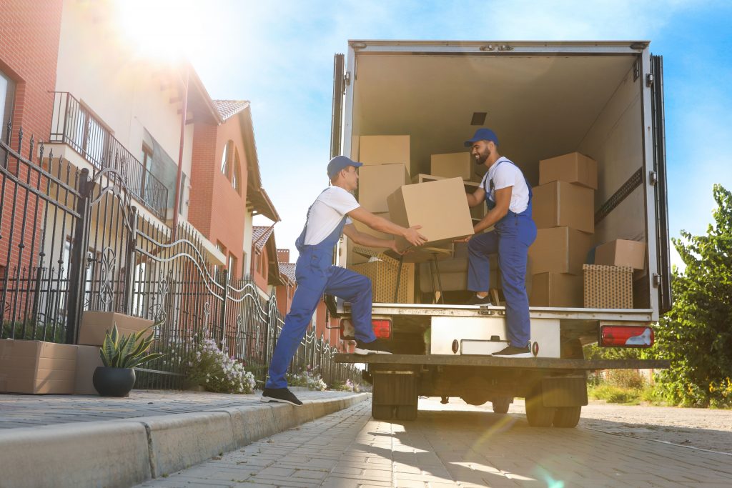 Two movers in blue uniforms are loading cardboard boxes into the back of a moving truck parked on a sunny residential street. Trusted movers in Lake & Orange County, FL ensure your belongings are safely transported to your new home.