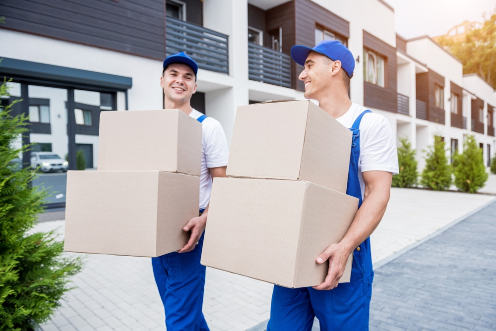 Two FL movers in blue uniforms and caps carry cardboard boxes, smiling at each other while walking outside a modern apartment complex on a sunny day—perfect service for Lake & Orange County.