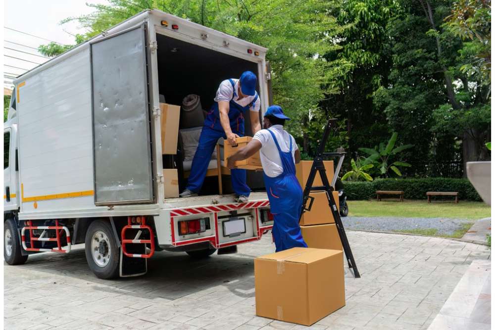 Two workers in blue uniforms from FL movers Lake & Orange County load cardboard boxes into the back of a moving truck parked on a driveway, surrounded by greenery and trees.