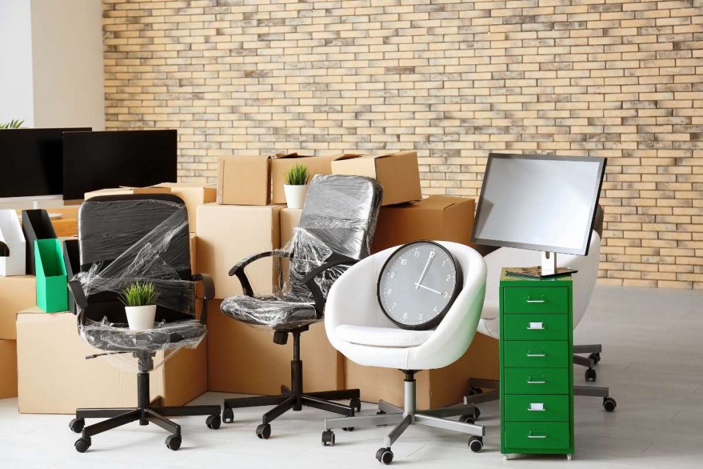 Office chairs, some wrapped in plastic, and boxes are stacked in front of a brick wall—ready for FL movers. A clock rests on a white chair. A green file cabinet with a computer monitor and potted plant is also visible. Perfect for movers Lake & Orange County.